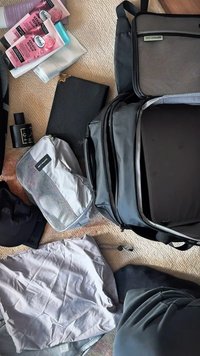 Szary plecak z siatkową kieszenią, otoczony różnymi przedmiotami, w tym kosmetykami w różowych tubkach, notesem oraz ubraniami w neutralnych kolorach.