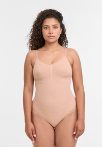 Žena s kudrnatými vlasy, oblečená v hladkém tělov�ém bodysuitu s tenkými nastavitelnými ramínky, stojící na jednobarevném bílém pozadí.