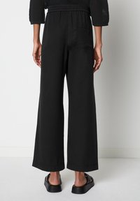 Zwarte wijde broek met een elastische tailleband, voorzien van zijzakken en een gladde structuur, gecombineerd met zwarte instap schoenen.