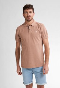 Hnědá bavlněná polo košile s límečkem, třemi knoflíky a malým logem na hrudi, kombinovaná s vyhrnutými světle modrými denimovými šortkami.