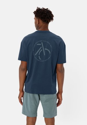 Camiseta de algodón azul marino con un gráfico circular de una bicicleta en la parte trasera, con el texto "BICICLETA DORMIR REPETIR" a su alrededor. Llevada con pantalones cortos verde claro.