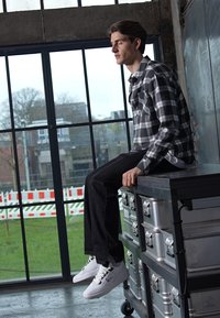 Mand siddende på en sølvfarvet opbevaringskasse, iført en sort og hvidternet skjorte, sorte jeans og hvide sneakers. Store vinduer i baggrunden.
