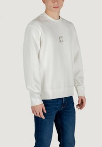 Weißes Sweatshirt mit geripptem Rundhalsausschnitt, langen Ärmeln und einem dezenten, grau bedruckten Logo auf der Brust, kombiniert mit blauen Jeans.