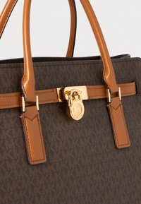 Sac à main en cuir marron avec des poignées en cuir beige, un fermoir en or et un motif "MK" répétitif sur le corps en tissu.