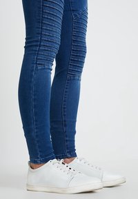 Modré skinny džíny s žebrovanými detaily na stehnech, spárovány s bílými nízkými teniskami. Hladká textura a přiléhavý design.