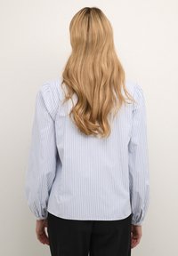 Blouse rayée présentant des rayures verticales bleu clair et blanches, des manches longues avec des poignets froncés, et un ourlet arrondi à l'arrière.