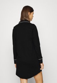 Vestido preto de mangas longas com um acabamento branco na gola e nos punhos, apresentando um corte reto e uma bainha arredondada na parte de trás.