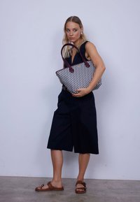 Torba na ramię z czarno-białym geometrycznym wzorem i bordowymi uchwytami. Modelka ma na sobie granatowy strój z szerokimi szortami i brązowymi sandałami.