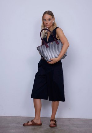 Sac en toile avec un motif géométrique noir et blanc et des poignées bordeaux. Le modèle porte une tenue marine composée de shorts à bas larges et de sandales marron.