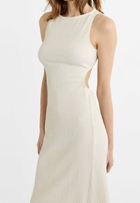 Robe sans manches en tricot écru avec un motif texturé en côtes et des découpes latérales, présentant un col haut et une silhouette ajustée.