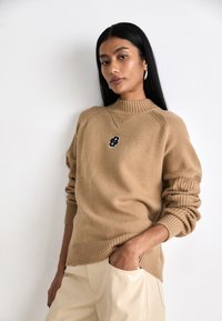 Sweater de malha bege com gola alta, punhos canelados e um detalhe de logótipo preto no peito. Combinado com calças de tom claro.