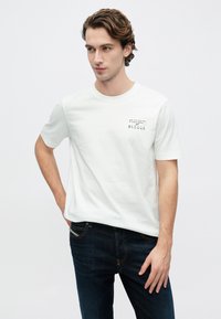 Bela bombažna majica s kratkimi rokavi, z črnim tiskanim logotipom na levi strani prsi. Kombinirana s temno modrimi denim kavbojkami.