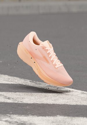 Roze sportschoen met dikke zool die boven een grijze weg met witte zebrapadstrepen en kleine vuildeeltjes eromheen zweeft.