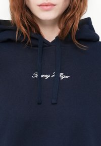 Marineblå hettegenser laget av bomull, med snor i hetta og hvit brodert "Tommy Hilfiger" logo på brystet. Glatt tekstur.