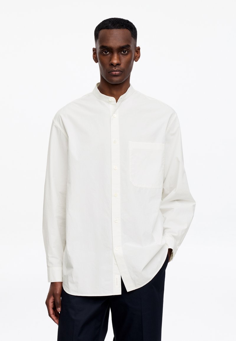 Jeune homme debout, portant une chemise blanche à col mandarin boutonnée et un pantalon sombre, sur un fond blanc uni.