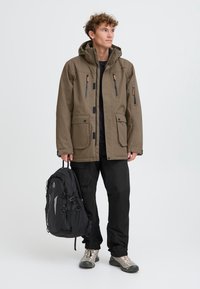 Olive roheline veekindel jakk koos kapuutsiga, ees taskud ja tõmblukuga detailid; koos mustade pükste ja musta seljakotiga. Hõbedased jalanõud.