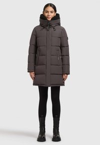 Abrigo acolchado en gris oscuro con cuello alto, cierre de cremallera, bolsillos laterales y diseño acolchado. Combinado con leggins negros y botas gruesas.