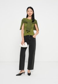 Groene top met een gedrapeerd ontwerp, zwarte rechte jeans en een witte geplooide clutch. Model draagt zwarte muilen, met een witte achtergrond.