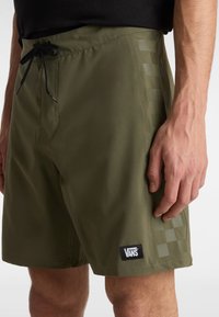 Shorts de bain olive avec une taille élastique à cordon de serrage, motif en damier sur le côté et un patch logo Vans noir près de l'ourlet.