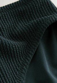 Sötétzöld szövet, amely texturált, bordás mintával és sima részekkel rendelkezik. Az anyag puha és rugalmas, a varrások jól láthatók.