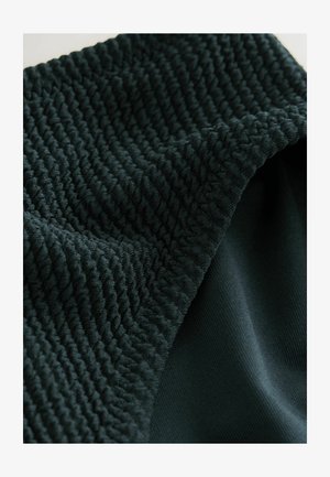 Sötétzöld szövet, amely texturált, bordás mintával és sima részekkel rendelkezik. Az anyag puha és rugalmas, a varrások jól láthatók.