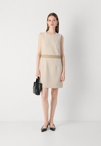 Beige mekanismilla varustettu hihaton mekko, jossa on tiukka vyötärö ja sileä vyö. Yhdistetty mustiin teräväkärkisiin korkokenkiin ja pieneen kudottuun käsilaukkuun.