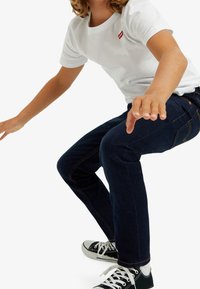 Blå denimjeans med gula sömmar, vit bomulls-T-shirt med röd logotyp och svarta kanvas sneakers. Person i en hukande pose.