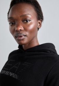 Crna hoodie s kapuljačom koja na prednjem dijelu ima teksturirani srebreni logo. Krupni plan tkanine i ovratnika, s glatkom završnom obradom.