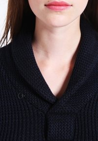 Námořnický svetr s texturou a šálovým límcem, vybavený velkým zapínáním na knoflík. Pletený vzor je silný a pohodlný, zdůrazňuje teplo.