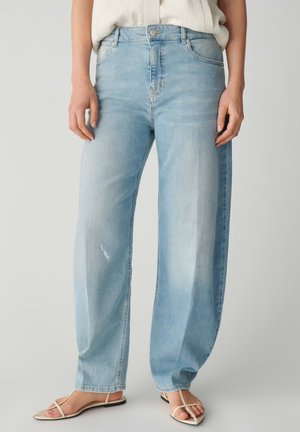 Lichtblauwe jeans met een losse pasvorm, gedragen met een wit shirt in de broek gestopt en beige platte sandalen met bandjes tegen een effen achtergrond.