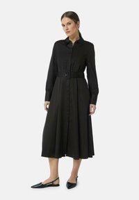 Schwarzes, langärmliges Hemdkleid mit Knopfleiste und breitem Gürtel. A-Linien-Silhouette mit glatter Textur. Kombiniert mit schwarzen spitzen Ballerinas.