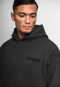 Czarna, oversize bluza z kapturem, wykonana z miękkiego materiału, z haftowanym na przedzie logo w ciemniejszym odcieniu, z kapturem na sznurku i luźnym krojem.