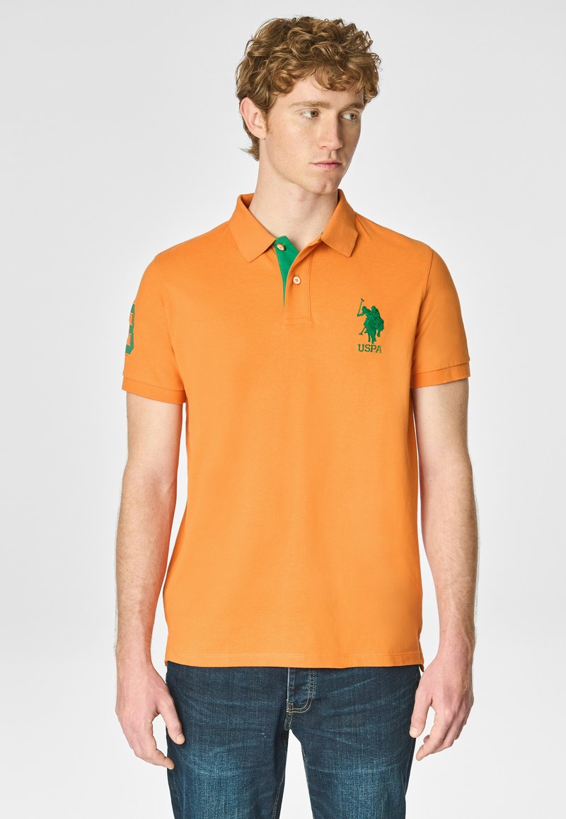 Polo arancione con colletto verde, dotato di logo ricamato verde e numero sulla manica. Realizzato in un tessuto liscio e traspirante.