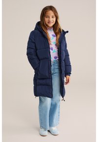 Mornarsko modra puhovka s kapuco, z edinstveno prešito zasnovo, z zadrgami in sproščenim krojem. Nosi se čez potiskano dolgo sleevel srajco in jeans.