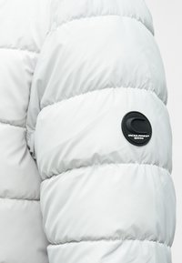 Ανοιχτό γκρι μπουφάν με στρώσεις, κατασκευασμένο από λείο, γυαλιστερό υλικό. Διαθέτει μαύρη στρογγυλή ετικέτα λογότυπου στο άνω μέρος του βραχίονα.