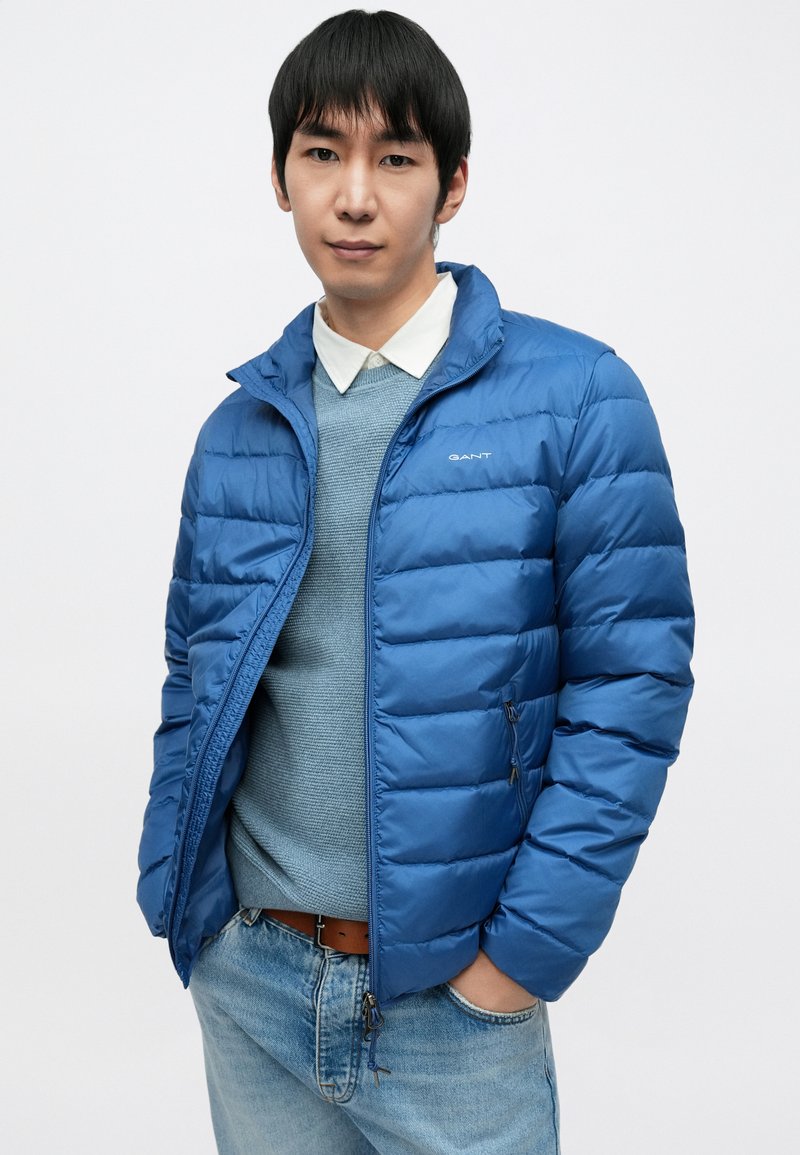 Blue puffer jacket with horizontal quilted design, zip closures, and high collar. Worn over a light blue sweater and paired with jeans.