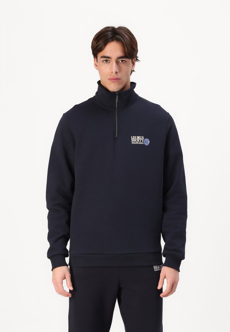 Felpa zip-up blu navy in un tessuto morbido, con colletto alto, polsini a coste e un logo sul petto per un tocco di dettaglio. Abbinata a pantaloni coordinati.