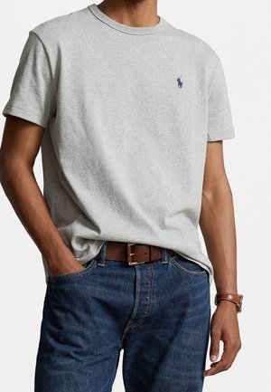 T-shirt gris à manches courtes avec un col rond et un logo bleu sur le côté gauche de la poitrine, associé à un jean en denim bleu et une ceinture marron.