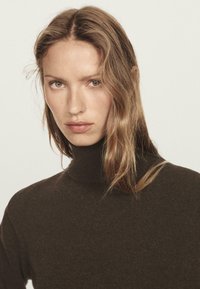 Frau mit hellbraunem Haar, die einen dunkelbraunen Rollkragenpullover trägt und direkt in die Kamera schaut, vor einem schlichten hellen Hintergrund.