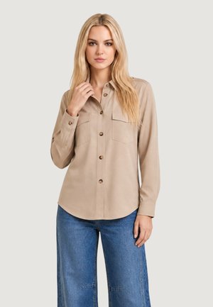 Donna con lunghi capelli biondi indossa una camicia beige con bottoni e tasche sul petto e jeans blu in denim a gamba larga su uno sfondo chiaro uniforme.