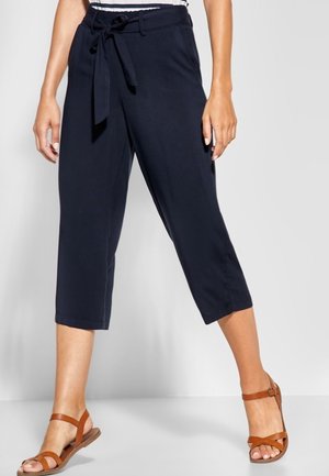 Frau trägt marineblaue, verkürzte Hosen mit gebundenem Taillengürtel und braune flache Sandalen, steht mit leicht gespreizten Beinen.