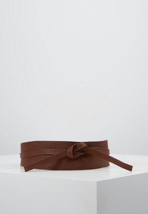 Ceinture large en cuir marron avec un design à double tour nouée en nœud simple, placée sur un piédestal blanc sur un fond uni.