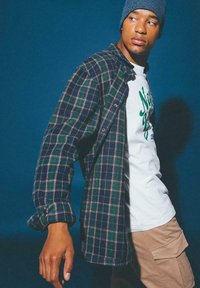 Koszula w kratę w kolorze granatowym, zielonym i różowym; noszona na białym T-shircie z zielonym napisem; beżowe spodnie cargo; niebieska dzianinowa czapka.