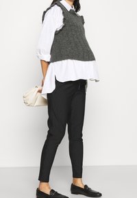 Chaleco de lana gris sobre una camisa blanca con cuello, combinado con pantalones negros a rayas y mocasines negros. Bolsa blanca en la mano.