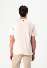 Helles rosa Baumwoll-T-Shirt mit kurzen Ärmeln, Rundhalsausschnitt und kleinem grünem Logo-Detail auf der Rückseite. Glatte Textur, lockere Passform.