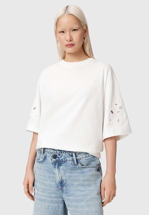 Jeune femme aux longs cheveux blonds portant une chemise blanche ample avec des découpes brodées sur les manches et un jean bleu clair.