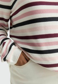 Close-up van een persoon die een geribbelde, gestreepte trui draagt in wit, roze, bordeauxrood en zwart, met een hand in de zak van lichtgekleurde broek.