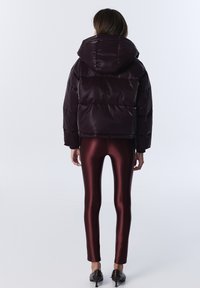 Doudoune en bordeaux profond avec capuche, présentant une finition brillante et un design matelassé, associée à des leggings bordeaux brillants et des chaussures noires.