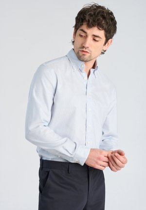 Uomo con camicia a righe azzurro chiaro con bottoni che si sistema i polsini, indossa pantaloni scuri, in piedi davanti a uno sfondo chiaro semplice.