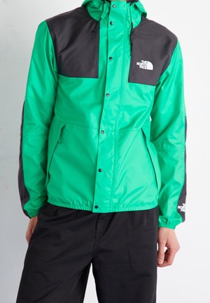 Hombre que lleva una chaqueta impermeable The North Face verde y negra con botones a presión y pantalones negros, de pie frente a un fondo liso.